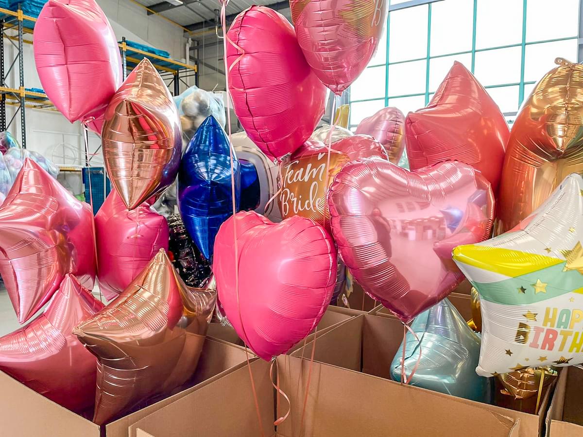 Colourful foil heart and star balloons for parties and events, including pink, rose gold, blue, and gold designs with ‘Team Bride’ and ‘Happy Birthday’ prints, displayed in warehouse